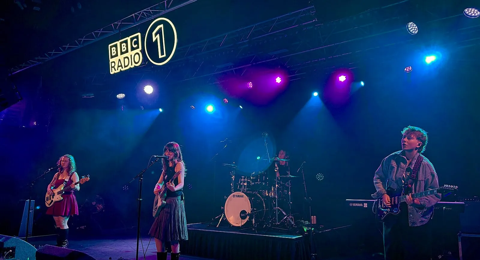 Florence Road’un dört üyesi mavi-mor ışıklı geniş bir sahnede, arkada BBC Radio 1 logosu