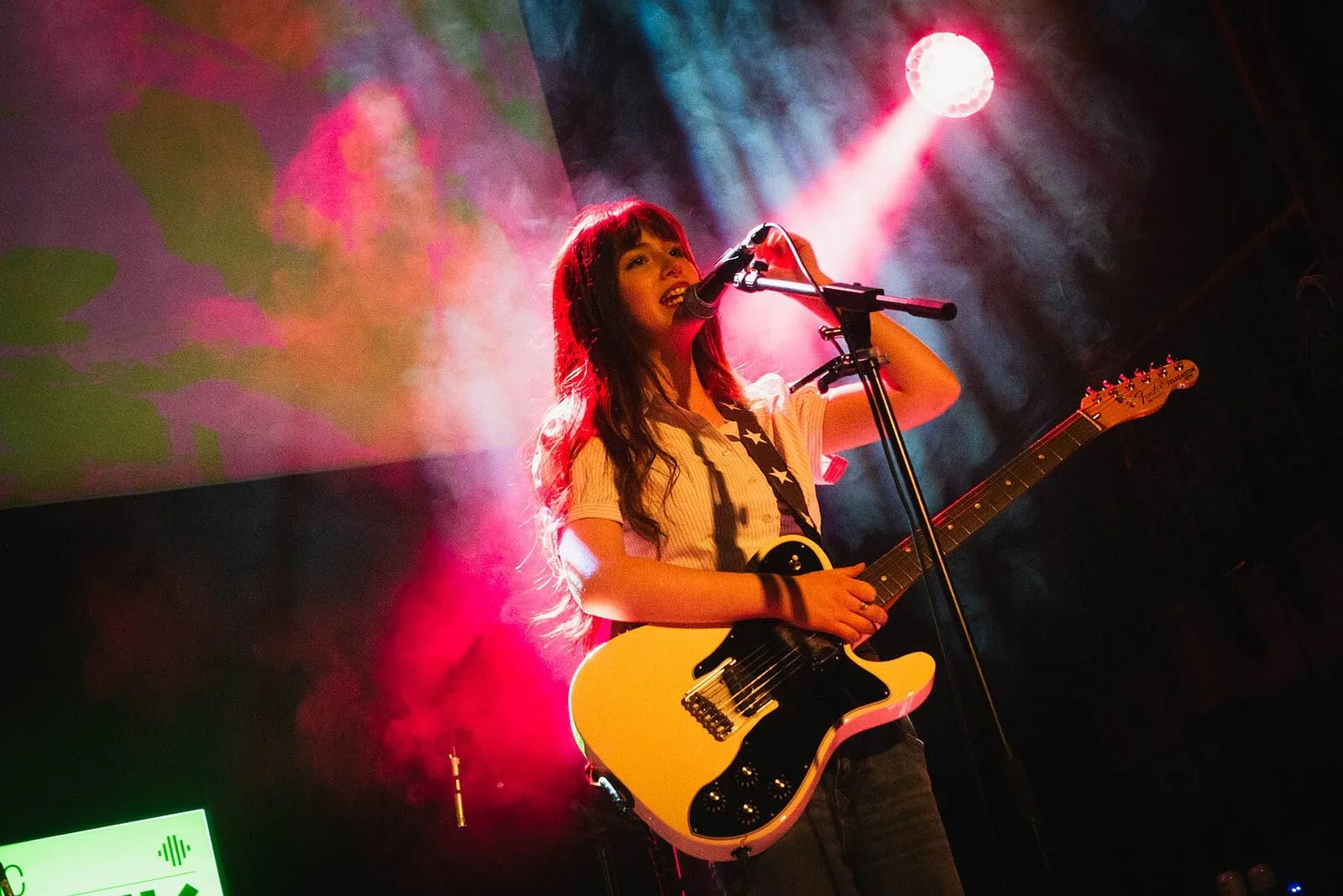 Lily Aron sahnede, pembe spot ışığı ve sis içinde, beyaz Telecaster’ıyla mikrofona eğilmiş